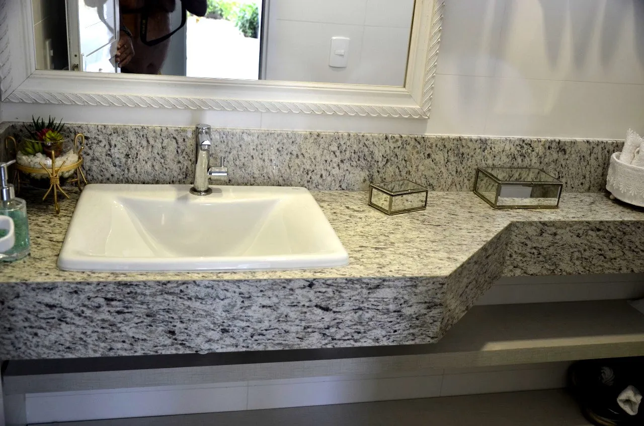 Bancada de granito claro com cuba embutida e saia lateral, em banheiro com box de vidro