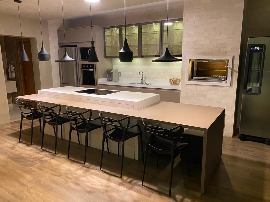 Bancada clara de área gourmet junto a mesa de jantar de madeira, com forno embutido ao fundo